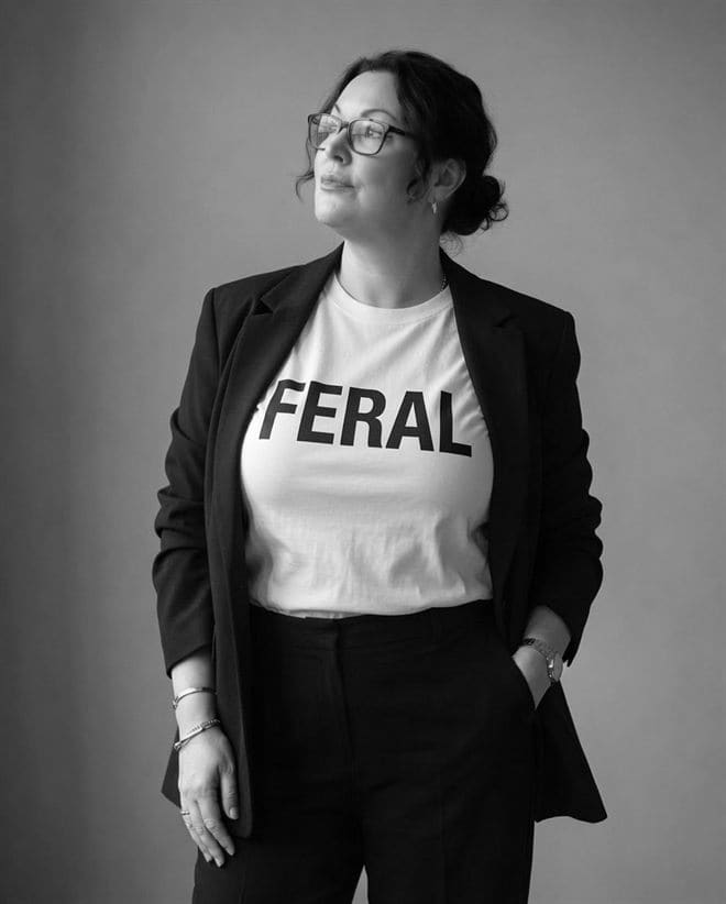 Sam Bearfoot, chin up and looking off camera, in a black blazer over a white FERAL T-shirt — a black-and-white studio portrait.